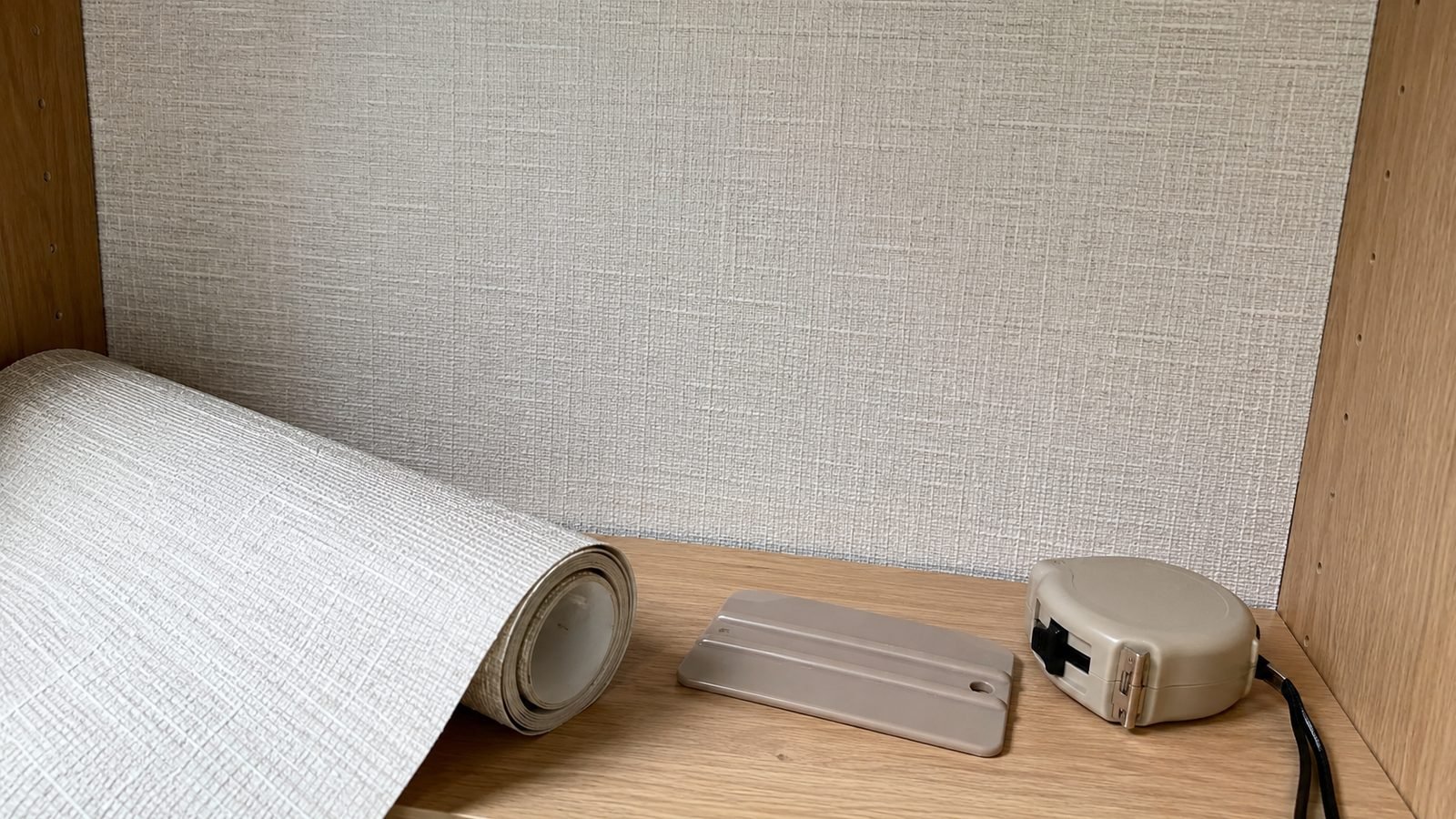 Close view of linen-texture peel-and-stick wallpaper on a small smooth panel with a plastic squeegee and tape measure nearby in soft daylight