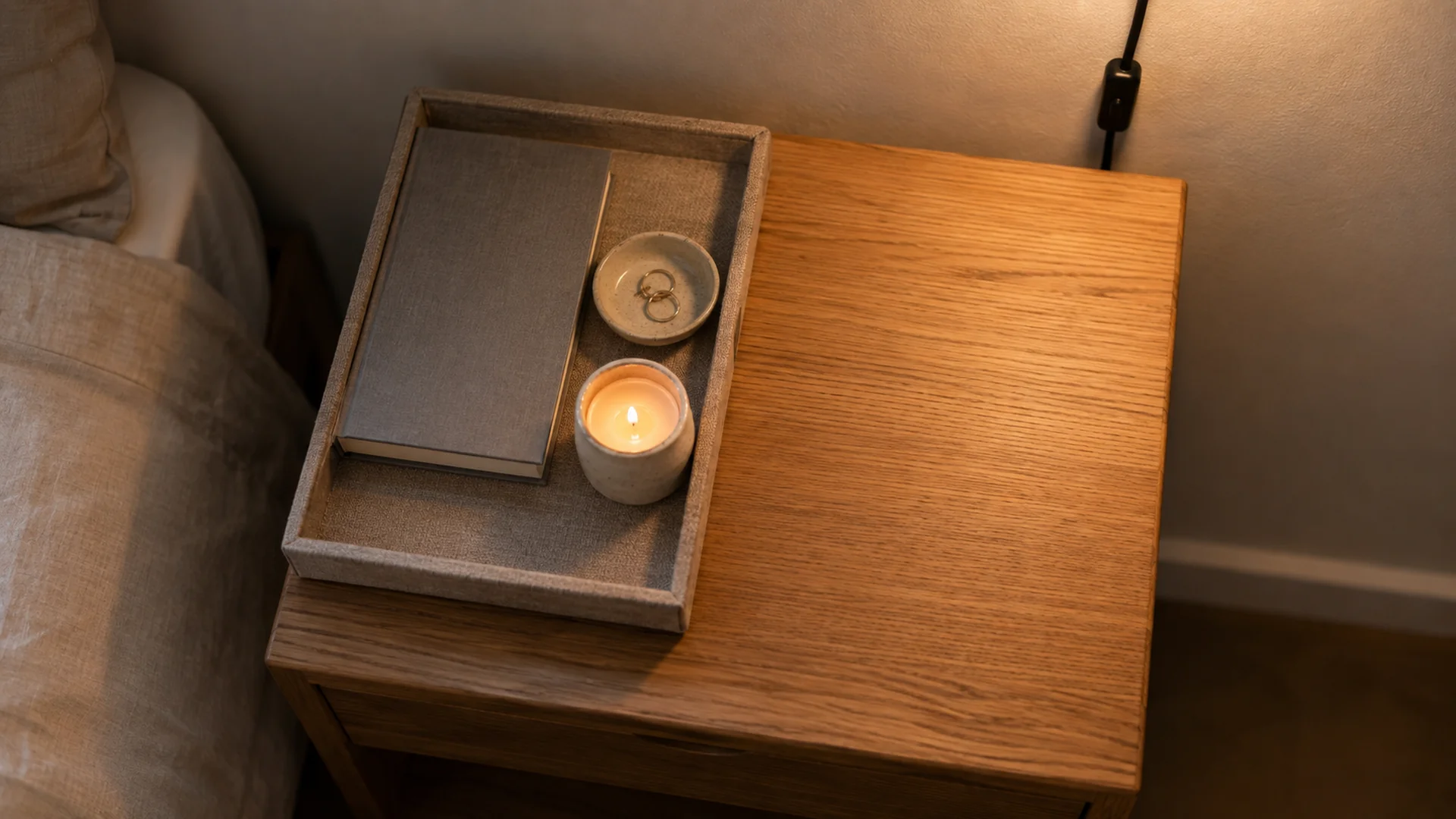Close-up of a tray on a tiny nightstand with one hardcover book, a small ceramic vase, and a neatly routed charging cord, plug-in sconce glowing warm 2700K above