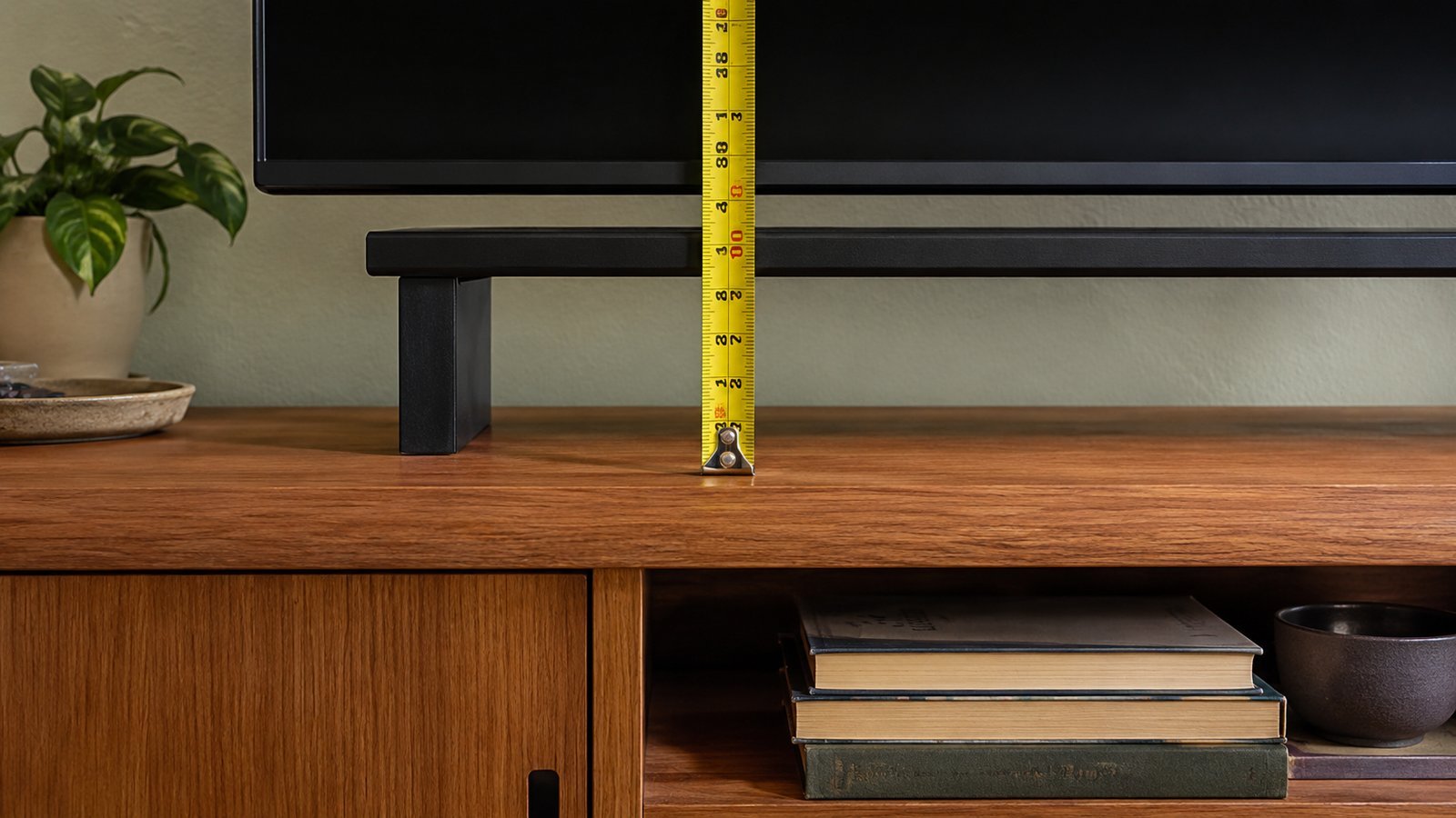 Yellow tape measure showing a 4–8 in gap between a wood console top and the bottom of a freestanding TV, cream sage rental wall, soft daylight